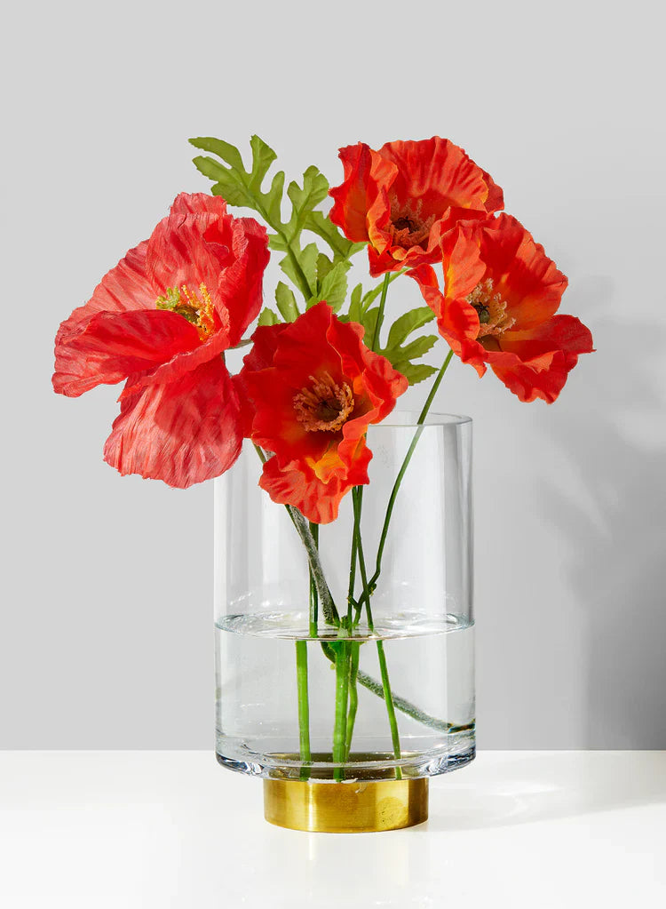 A clear vase with water holds six vibrant orange-red calla lilies, set on a wooden surface against a neutral, textured background.