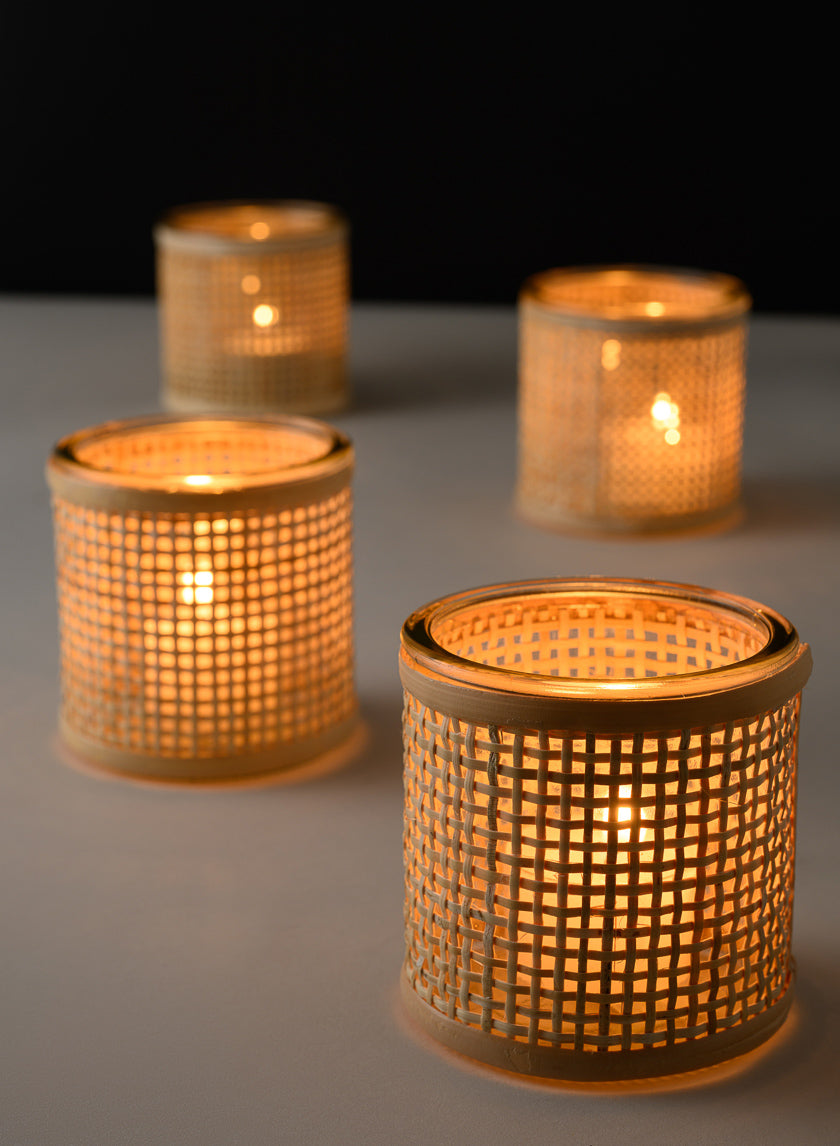 Four glass candle holders wrapped in woven rattan, each containing a lit tealight candle, are arranged on a light surface against a dark background, casting a warm, cozy glow.